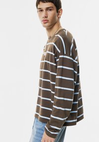 Jongeman in een bruin en lichtblauw gestreept overhemd met lange mouwen en lichtblauwe jeans, staand tegen een witte achtergrond, iets zijwaarts gekeerd.