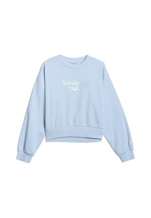 Młoda dziewczynka stoi, ubrana w jasnoniebieski komplet bluzy i spodni dresowych z napisem "Winter Vibes" oraz dwukolorowe fioletowo-białe trampki.