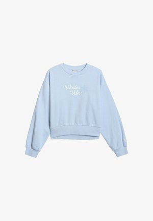 Młoda dziewczynka stoi, ubrana w jasnoniebieski komplet bluzy i spodni dresowych z napisem "Winter Vibes" oraz dwukolorowe fioletowo-białe trampki.