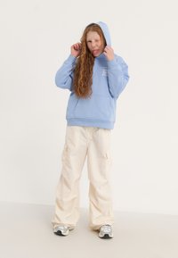 Gai zili pelēkā kapu žakete ar priekšējo kabatu, savienota ar krēmkrāsas kravu biksēm. Modelis nes pelēkas kedas un viņai ir garas, viļņotas sarkanas matu cirtas.