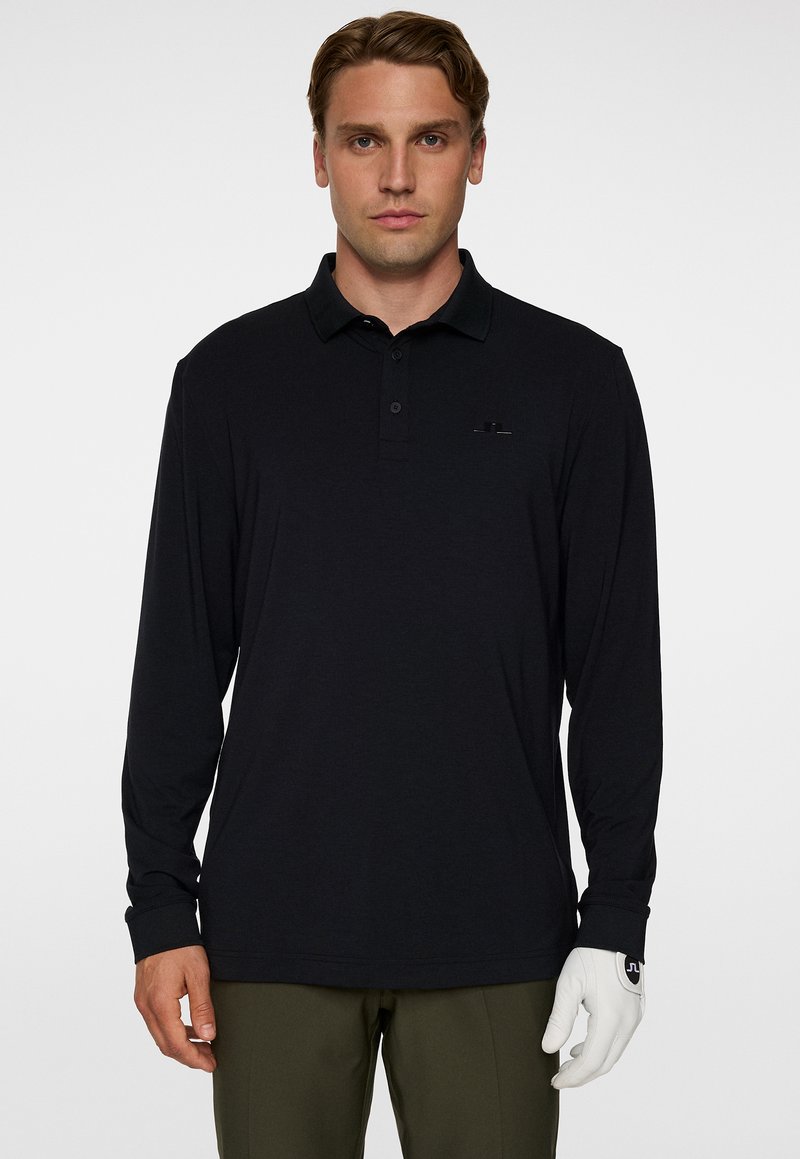 Mann trägt ein schwarzes Langarm-Polohemd und olivfarbene Hosen, mit einem weißen Golfschuh an seiner linken Hand, steht vor einem weißen Hintergrund.