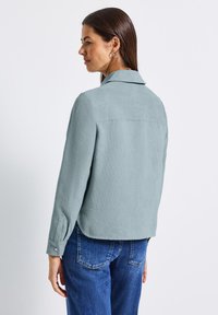 Světle modrá, texturovaná blůza s límečkem, dlouhými rukávy a knoflíkovými manžetami. Má zakřivený lem a detail zadní části. V kombinaci s modrými džíny.