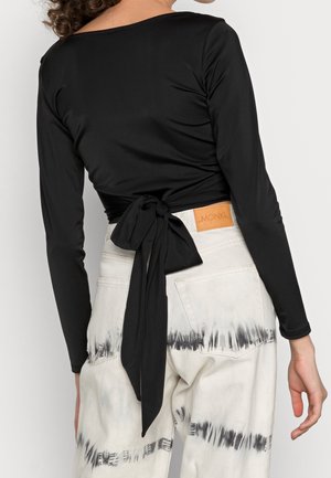 Kvinde iført sort langærmet slå-om top bundet bagpå, kombineret med hvide højtaljede jeans med sort tie-dye og baglommer.
