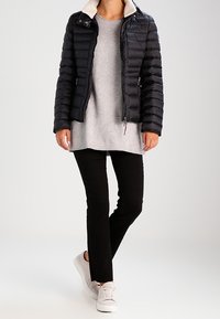 Doudoune noire avec un matelassage horizontal et un col en fausse peau de mouton clair, portée sur un haut gris et un pantalon noir, complétée par des baskets blanches.