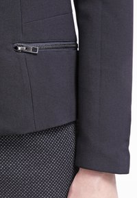 Czarna dopasowana marynarka z detalem w postaci kieszeni na zamek, gładką teksturą tkaniny i zorganizowanym kształtem. Nosi się ją na wzorzystą szaro-różową spódnicę.