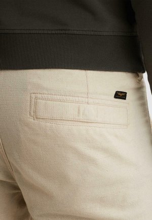 Pantalones beige con textura con una pequeña etiqueta de marca negra y dorada cerca de un bolsillo trasero de ribete, combinados con una manga y bajo de suéter oscuro con canalé.