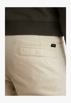 Pantalones beige con textura con una pequeña etiqueta de marca negra y dorada cerca de un bolsillo trasero de ribete, combinados con una manga y bajo de suéter oscuro con canalé.