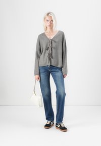 Γκρι πλεκτό πουλόβερ με V λαιμόκοψη και διακοσμητικές κορδέλες, συνδυασμένο με ανοιχτό μπλε τζιν. Κρατάει μια λευκή τσάντα, φοράει μαύρα και καφέ αθλητικά παπούτσια.