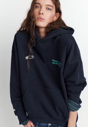 Navyblå hættetrøje med en forlompe, der har en hvid logo og grøn tekst, der lyder "Følg drømme, ikke likes." Lange ærmer med ternede manchetter.