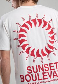 White cotton t-shirt featuring a large red sun graphic on the back and bold red text reading "SUNSET BOULEVARD" at the bottom.