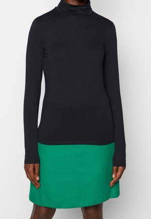 Osoba ubrana w czarny golf z długim rękawem, wpuszczony w jasną zieloną spódnicę, stojąca na tle jednolitego białego tła.