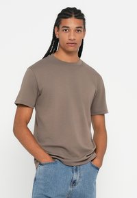 Νεαρός άντρας με μακριά πλεχτά μαλλιά, ντυμένος με ένα απλό καφέ κοντομάνικο μπλουζάκι και ανοιχτόχρωμα μπλε τζιν, στέκεται με τα χέρια στις τσέπες.