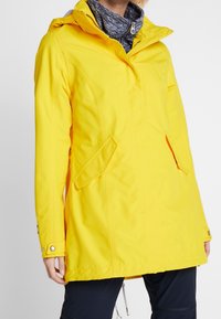 Kobieta w jasnoniebieskim długim płaszczu przeciwdeszczowym z kapturem, na granatowej bluzie zapinanej na zamek i czarnych spodniach, na jednolitym tle.