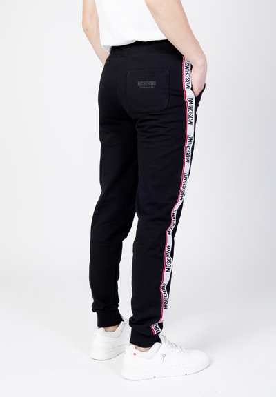 Czarne dresy Moschino z białą i czerwoną taśmą logo po bokach. Posiadają elastyczny pas, ściągacze na kostkach oraz tylną kieszeń z detalem logo.