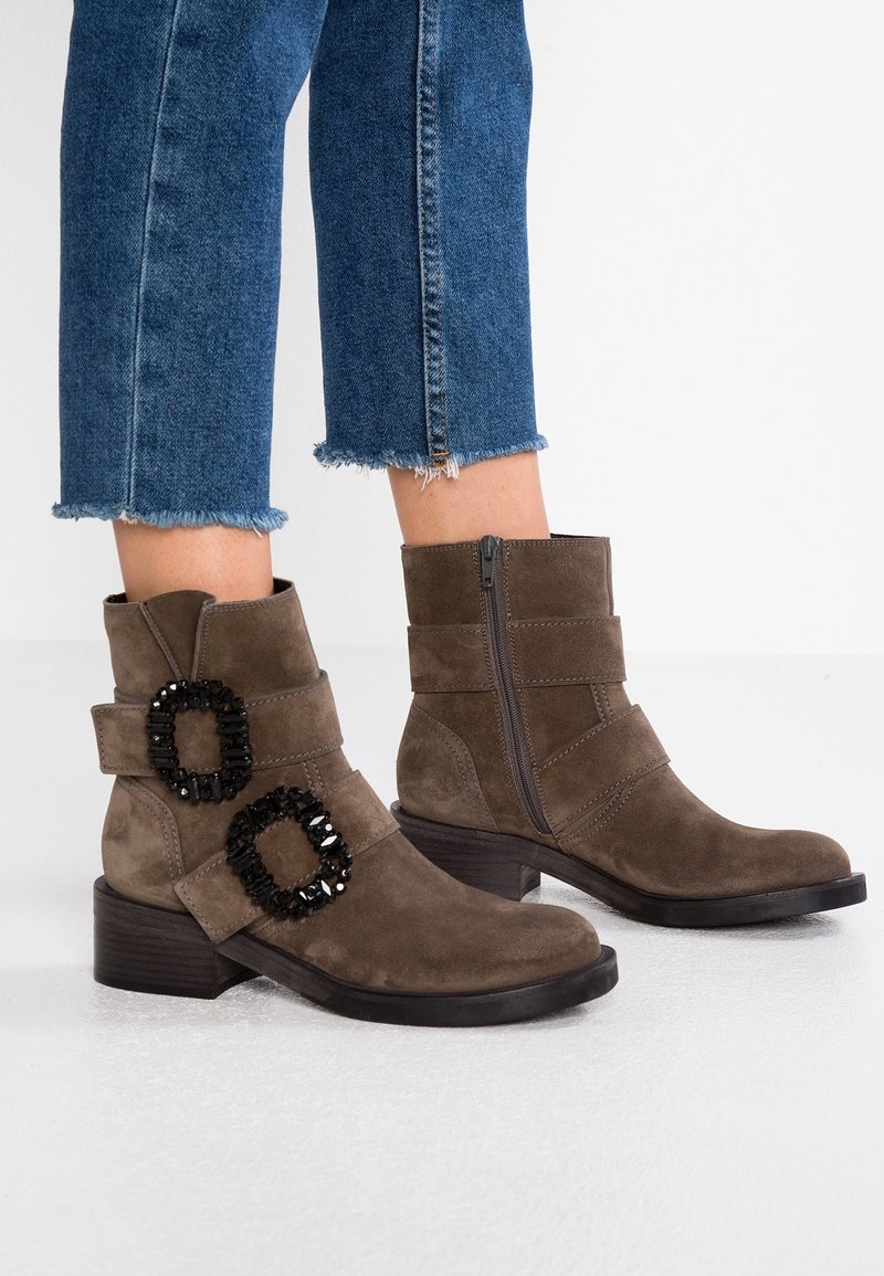 Bottines en daim marron avec des brides à boucle ornées de perles noires, fermeture éclair latérale, talon bas empilé, portées avec un jean en denim bleu à ourlet effiloché.