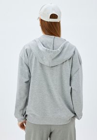 Sudadera con capucha gris hecha de una tela suave, con un corte holgado, puños acanalados y una costura centrada en la parte posterior. Se lleva con una gorra de color claro.