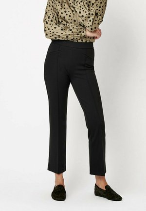 Schwarze, maßgeschneiderte Hose mit geradem Schnitt, ausgestattet mit Vordertaschen und einer glatten Textur, kombiniert mit einer beigen Bluse mit Punkten und dunklen Schuhen.