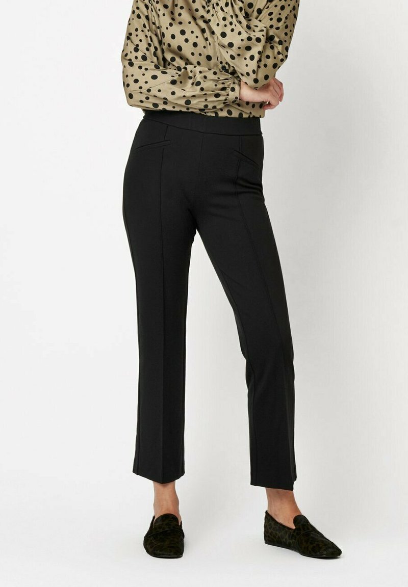 Czarne spodnie o prostym kroju, z kieszeniami z przodu i gładką teksturą, zestawione z beżową bluzką w kropki oraz ciemnymi butami.