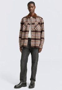 Flanellhemd in braun, creme und rot kariert. Verfügt über zwei Brusttaschen und einen kontrastierenden braunen Kragen. Kombiniert mit schwarzer Jeans und Stiefeln.