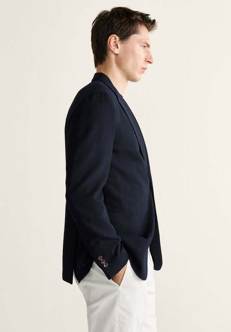 Hombre con blazer azul marino y pantalones blancos de perfil con las manos en los bolsillos sobre un fondo liso.