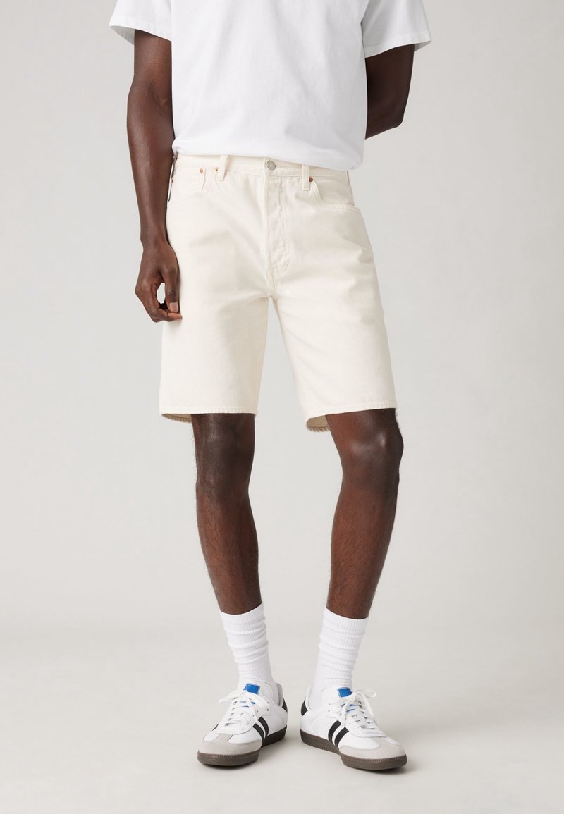 Homme portant un short en jean blanc cassé, des chaussettes blanches montantes, des baskets blanches avec des rayures noires, et une chemise blanche à manches courtes, debout devant un fond uni.