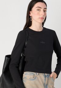 Model s dugom tamnom kosom nosi crni duksericu dugih rukava. Nosii veliku crnu torbu i ima srebrne kružne naušnice. Minimalistička pozadina.