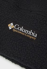 Čierna fleecová látka s textúrovaným povrchom. Obsahuje obdĺžnikovú nálepku s nápisom "Columbia Sportswear Company" v bielej a zlatej farbe.