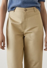 Beżowe dopasowane spodnie o gładkiej fakturze, z doszytymi przednimi kieszeniami i pojedynczym zapięciem na guzik w talii. W zestawie z niebieskim swetrem dzianinowym.