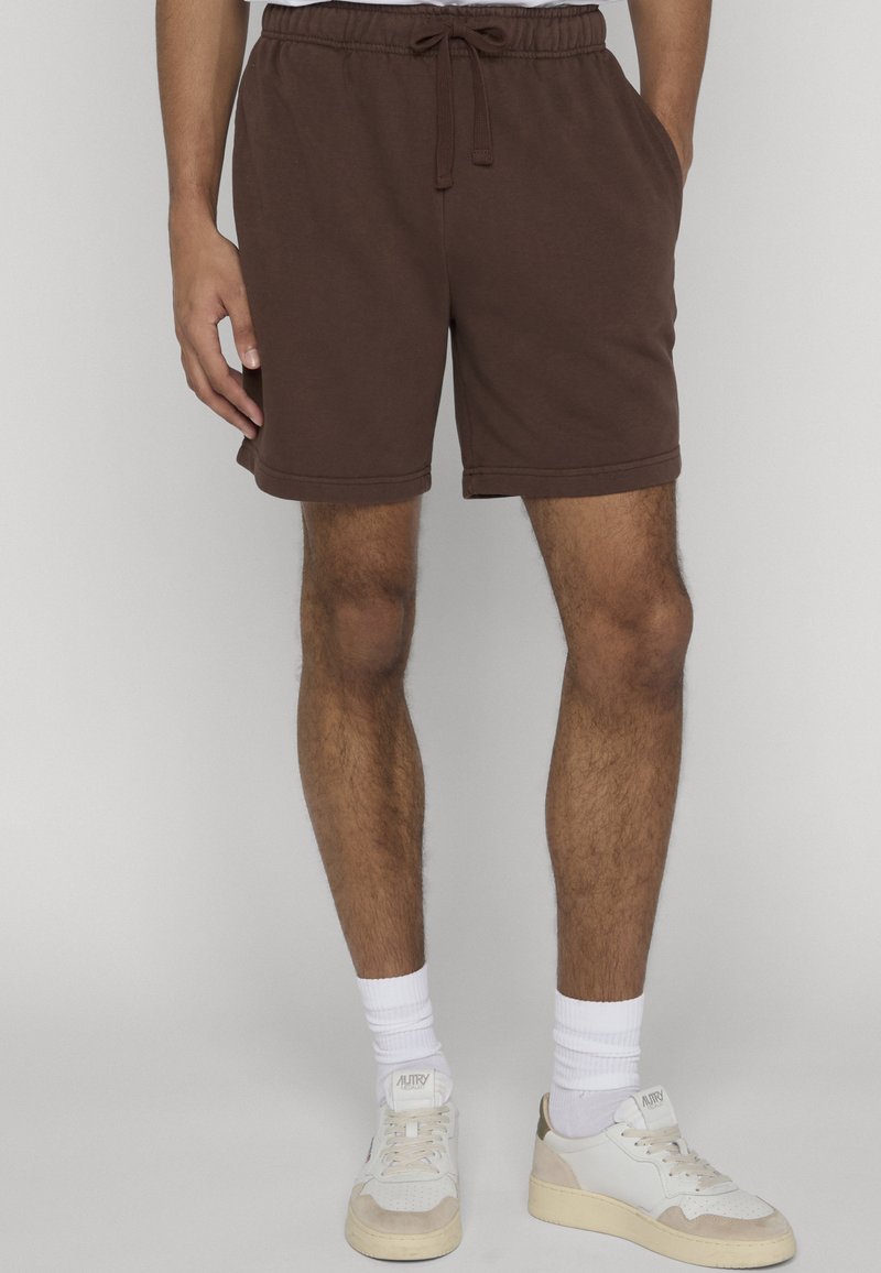 Homme portant un short à cordon de serrage marron, des chaussettes blanches et des baskets blanches, debout avec une main dans la poche devant un fond uni.