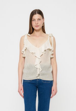 Vrouw met steil bruin haar die een crèmekleurige mouwloze blouse met ruches en strikbandjes draagt en blauwe spijkerbroek, staand tegen een effen witte achtergrond.