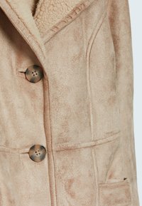 Tan faux suède jas met een gestructureerde afwerking, gekartelde kraag, twee voorkruiszakken en bruine knopen. Ontwerp bevat zichtbare stikseldetails.