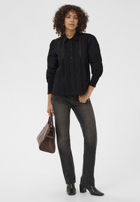 Femme portant un chemisier noir à boutons et un jean foncé, tenant un sac à main marron, se tenant devant un fond clair.