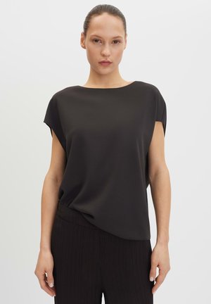 Frau mit zurückgebundenem Haar, die ein locker sitzendes schwarzes ärmelloses Oberteil und schwarze Hose vor einem einfarbigen hellen Hintergrund trägt.