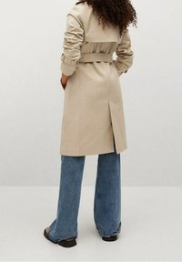 Beżowy trencz z paskiem w talii, naramiennikami i rozcięciem na plecach, w połączeniu z niebieskimi dzwonami i czarnymi sandałami.