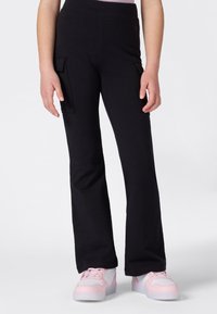 Personne portant un pantalon cargo noir évasé et des baskets blanches avec des accents roses, se tenant contre un fond clair et uni.