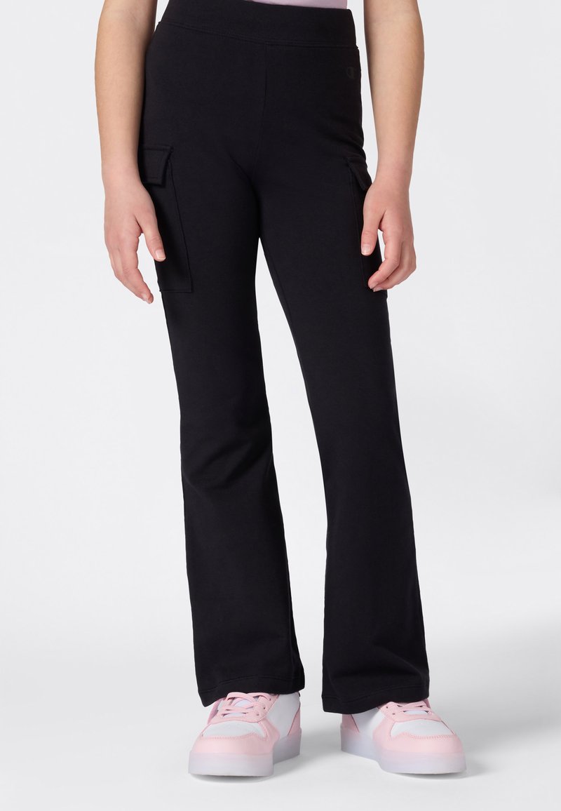 Personne portant un pantalon cargo noir évasé et des baskets blanches avec des accents roses, se tenant contre un fond clair et uni.
