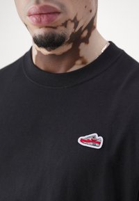 Svart bomull t-shirt med en liten broderad röd sneakers-logo på bröstet. Materialet verkar mjukt med en klassisk rundad halsringning.