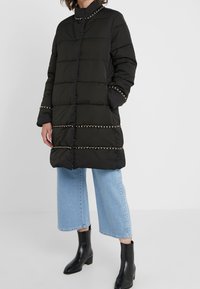 Czarna puchowa kurtka z przeszyciami w poziome pasy, metalicznymi łańcuszkami przy kołnierzu i na dole oraz zapinana na guziki. Nosi się ją na jasne dżinsy.