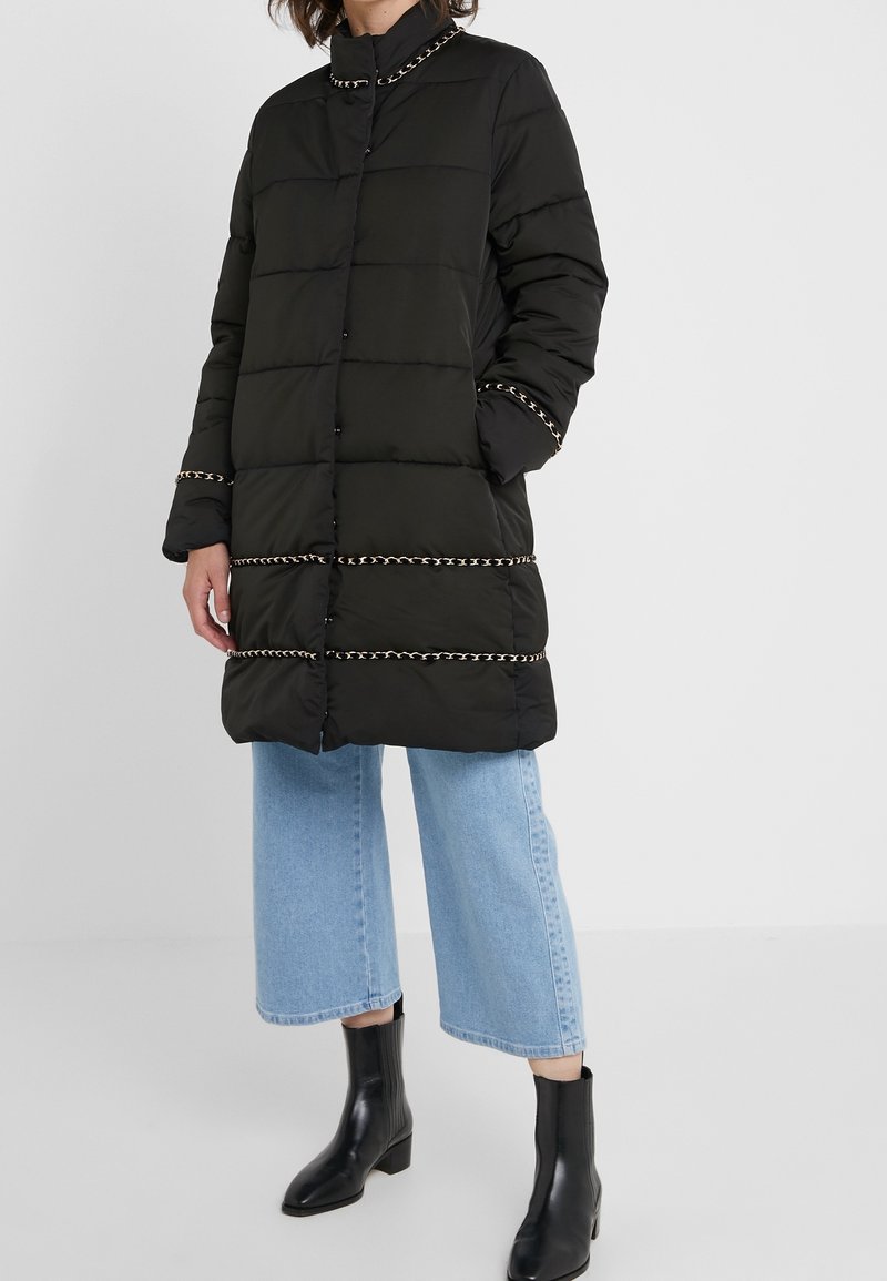 Czarna puchowa kurtka z przeszyciami w poziome pasy, metalicznymi łańcuszkami przy kołnierzu i na dole oraz zapinana na guziki. Nosi się ją na jasne dżinsy.