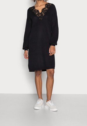 Abito lavorato a maglia nero con scollo a V e dettagli in pizzo intricati. Maniche lunghe e vestibilità comoda, abbinato a sneaker bianche.