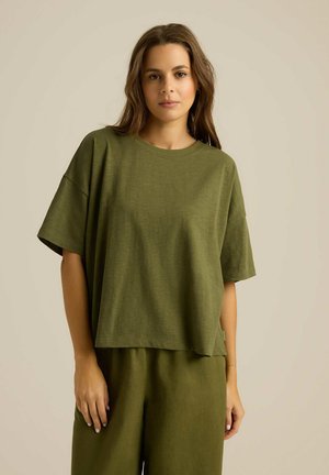Oliwkowy top z krótkim rękawem o luźnym kroju, wykonany z miękkiej tkaniny. Posiada okrągły dekolt i zaokrąglony dół, w zestawie ze pasującymi spodniami.