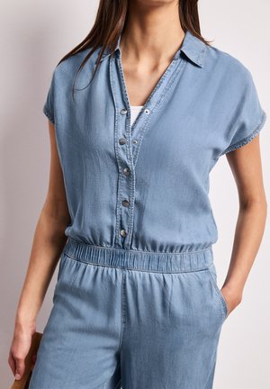 Femme portant une combinaison à manches courtes bleu clair avec boutons, taille élastique et mains dans les poches, sur fond uni.