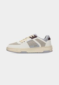 Baskets basses beige, gris et blanc avec des lacets jaunes et le logo "HUGO" sur le côté, sur un fond clair uni.