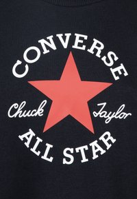 Black fabric with a large red star in the center, featuring white text that reads "CONVERSE" above and "ALL STAR" below, and "Chuck Taylor" in cursive.