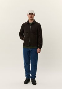 Ruda striukė su užtrauktuku ir dviem priekinėmis kišenėmis, dėvima ant žalios marškinėlių, derinama su mėlynais džinsais ir juodais batais. Šviesios spalvos kepurė papildo įvaizdį.