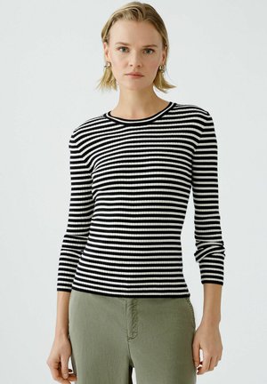 Haut à manches longues rayé noir et blanc en côtes, avec un col rond, présentant un design ajusté et un tissu texturé. Associé à un pantalon vert.