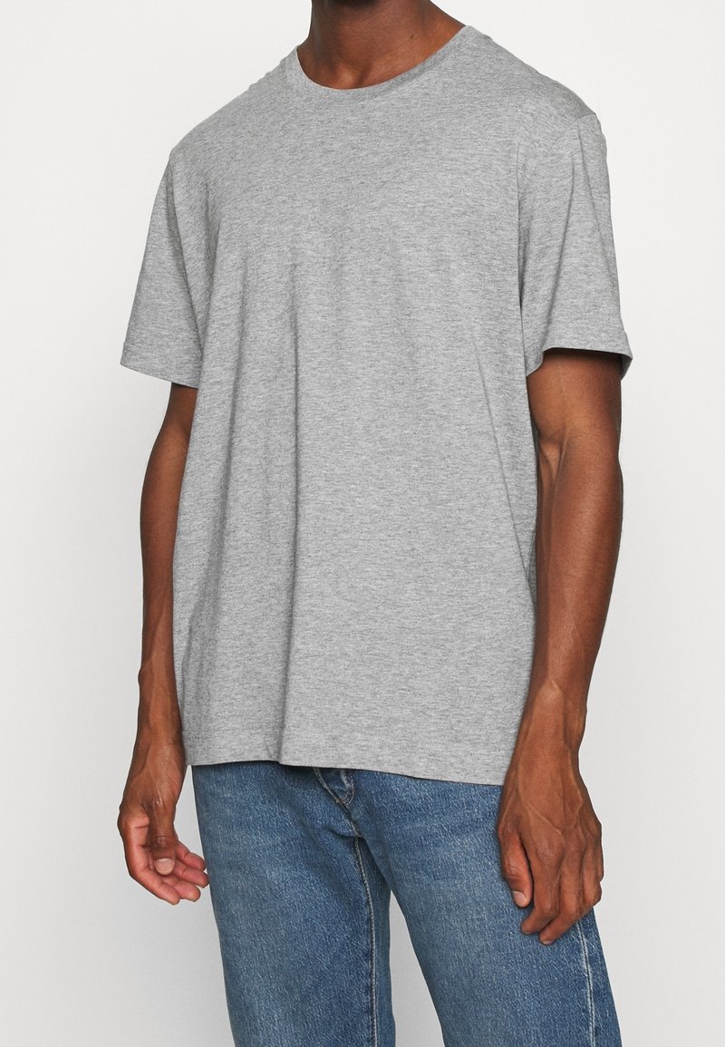Homme portant un t-shirt gris uni à manches courtes et un jean bleu, debout devant un fond blanc uni.