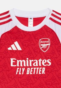 Röd Adidas-tröja med vita detaljer, prydd med Arsenal-logotyp och texten "Emirates Fly Better". Materialet har ett subtilt mönster.