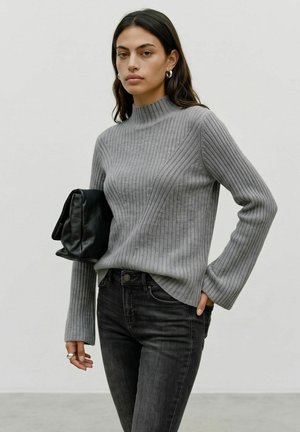 Femme portant un pull côtelé gris et un jean noir, tenant une pochette en cuir noire sous le bras sur un fond uni.