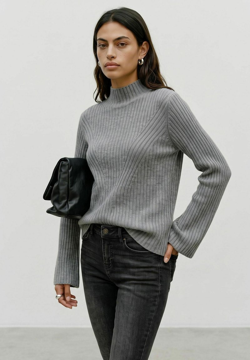 Femme portant un pull côtelé gris et un jean noir, tenant une pochette en cuir noire sous le bras sur un fond uni.